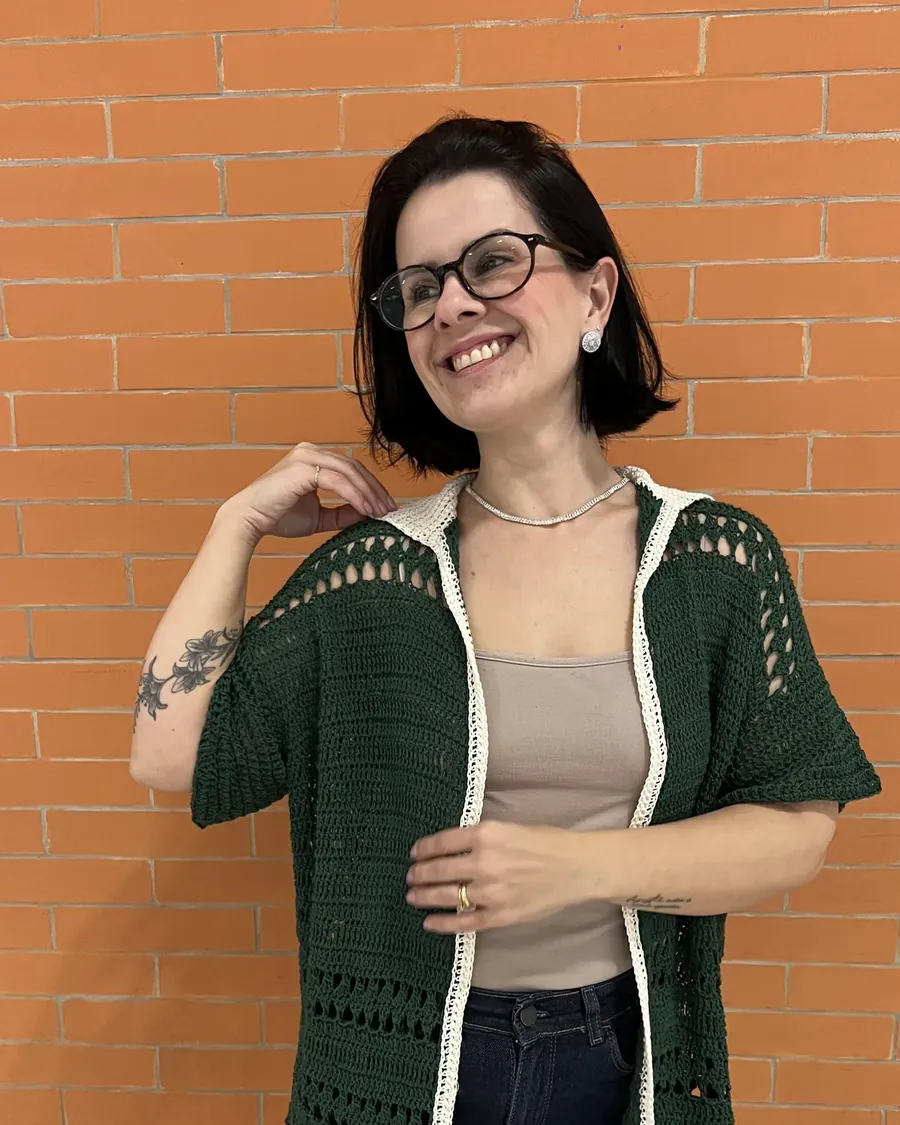 Gabi Silva sorrindo com um casaco verde de crochê feito por ela, em frente a uma parede de tijolos laranja