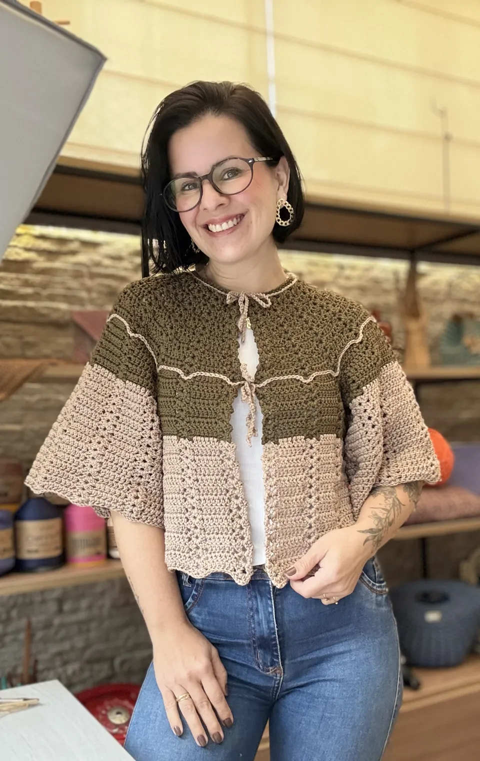 Gabi Silva sorrindo para a câmera no ateliê, vestindo um casaquinho de crochê feito por ela, com fios de crochê na estante ao fundo
